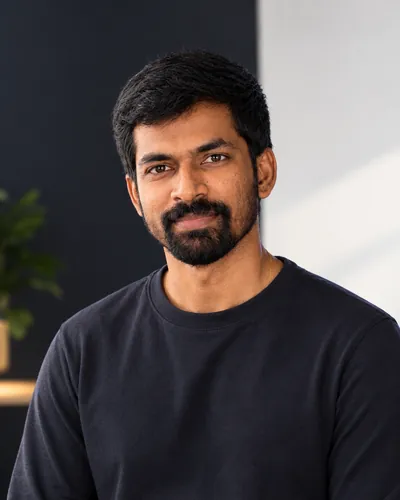 Portrait of Haribaskar Dhanabalan smiling, in a dark t-shirt, in a bright room with a plant behind him